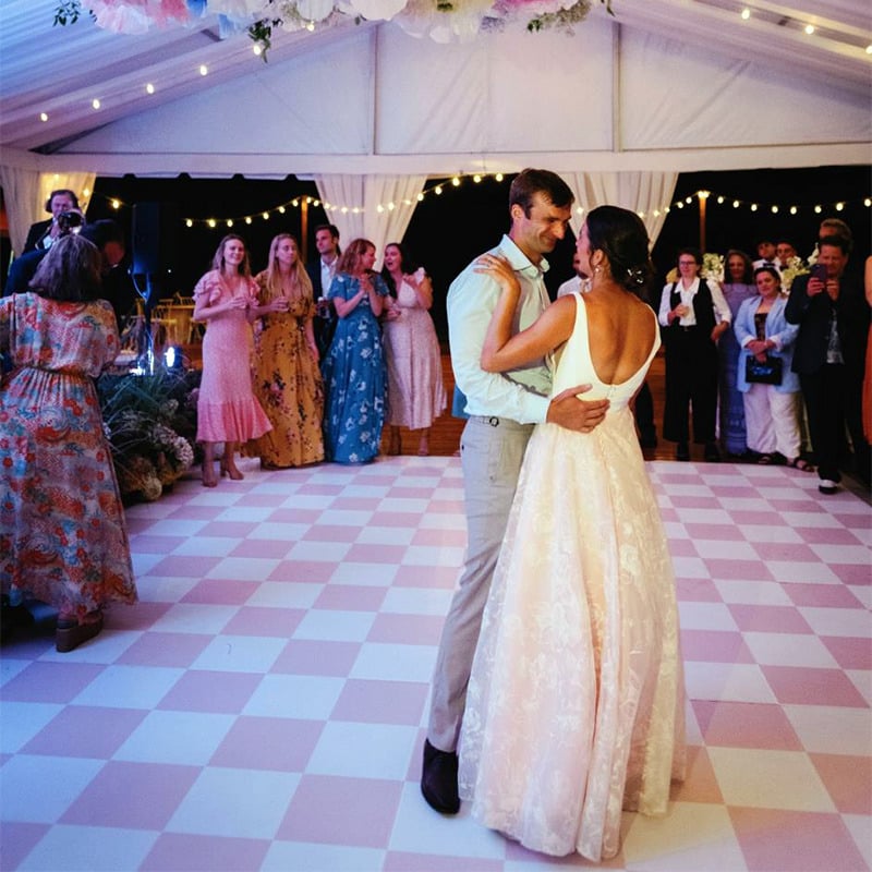 A bride and groom dance upon a rented dance floor that's been covered with a pink and white checkered vinyl dance floor wrap at their outside wedding under a big, white tent.