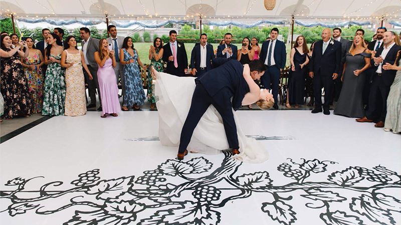 A design detail of a grapevine, taken from the embroidery on the bride's dress, has been printed on the vinyl dance floor wrap covering the rented dance floor at this outside wedding.