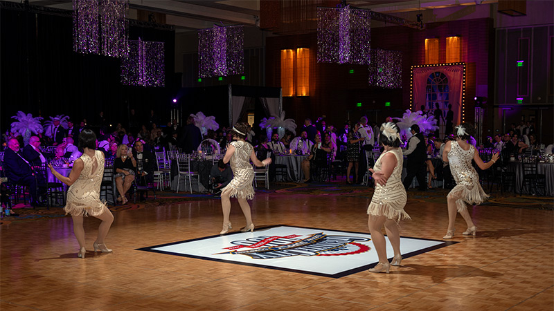 Dancers performing a jazz routine do so upon a vinyl dance floor wrap that has been custom printed with the corporate name and logo at a company gathering and celebration event.