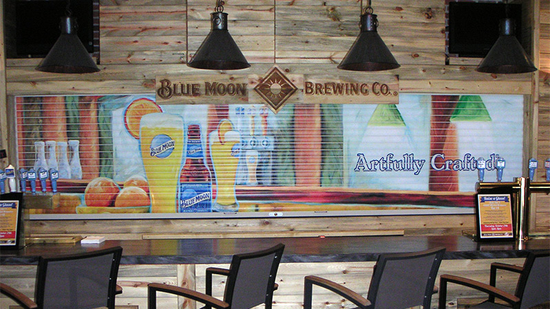 A Blue Moon Pale Ale branded wallpaper mural covers the corrugated metal enclosure keeping the beer taps safe at The Sandlot Brewery inside Coors Field in Denver, Colorado.