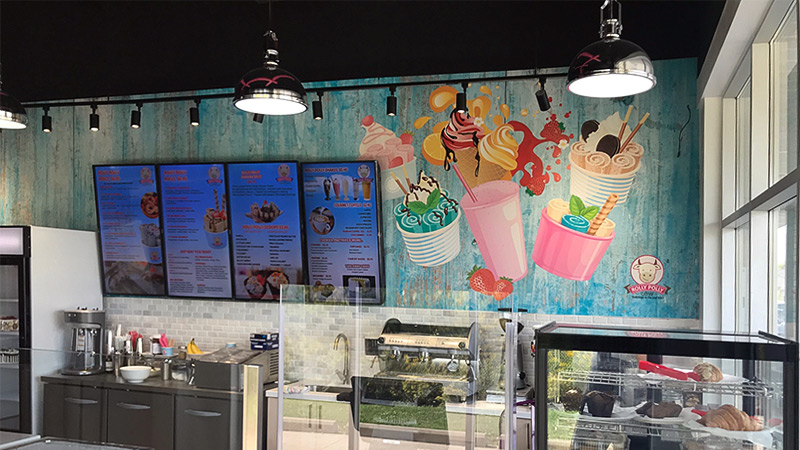 Customers at Rolly Polly Cow Ice Cream in Brampton, Ontario are greeted by a custom designed wallpaper mural covering the entire back wall of the service counter. This signage at the franchise includes whimsical ice cream shakes and cones of ice cream.