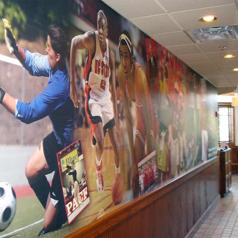 A wallpaper mural featuring a collage of local high school and college athletes lines both sides of the hallway at Backyard Bistro, and American restaurant and sports bar in Raleigh, NC. It's a great way to show community pride.