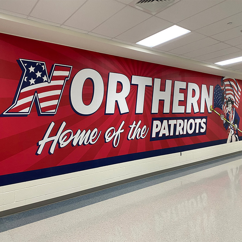 A large printed wallpaper mural installed on a cinder block wall in this school hallway welcomes students and visitors with the school&rsquo;s mascot, team name and school colors.
