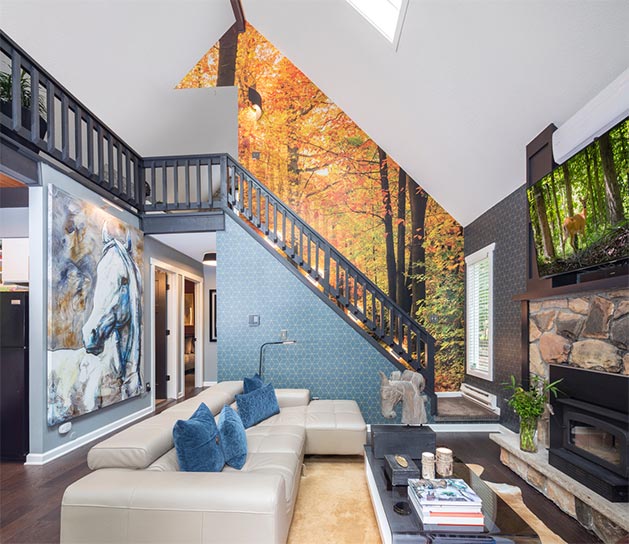 living room with cream couch, geometric pattern wallpaper accent walls and colorful fall foliage forest wall mural going up stairwell's vaulted ceiling