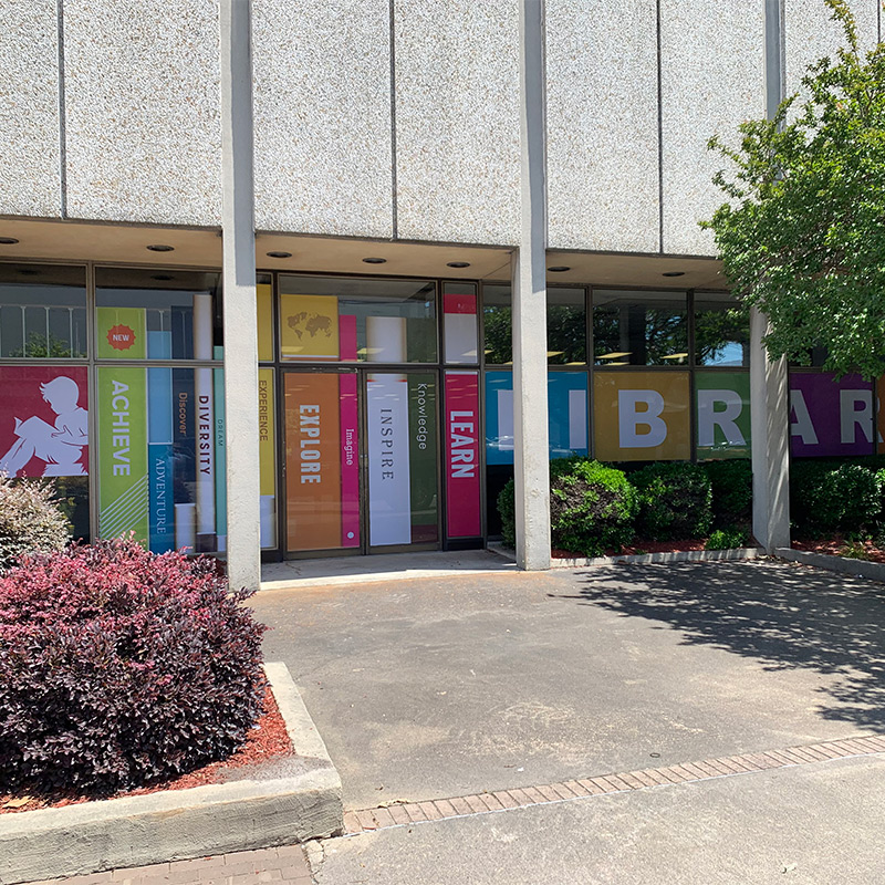 Colorful perforated window graphics cover the exterior glass of a commercial looking building that houses a library.
